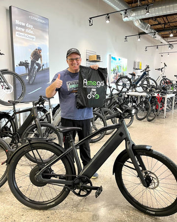 A man in a bike shop stands behind a matte black e-bike, holding up a black branded Amego tote bag. The shop is filled with other bicycles.