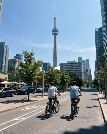 Two cyclists ride on a city street toward the Toronto CN Tower, with tall buildings, trees, parked cars, and a clear blue sky in the background.